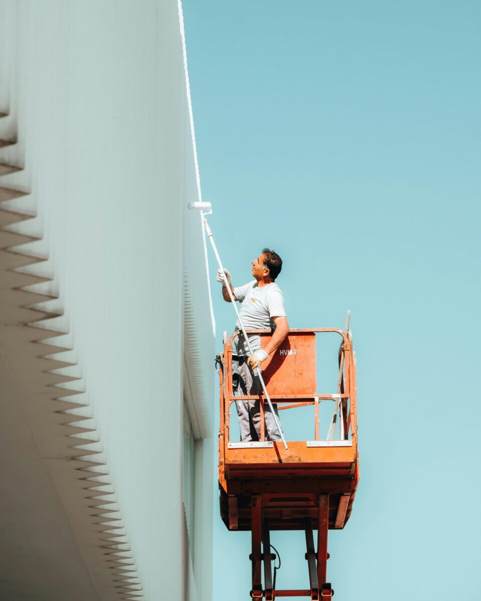 A man painting a building exterior from a cherry picker against a clear blue sky.